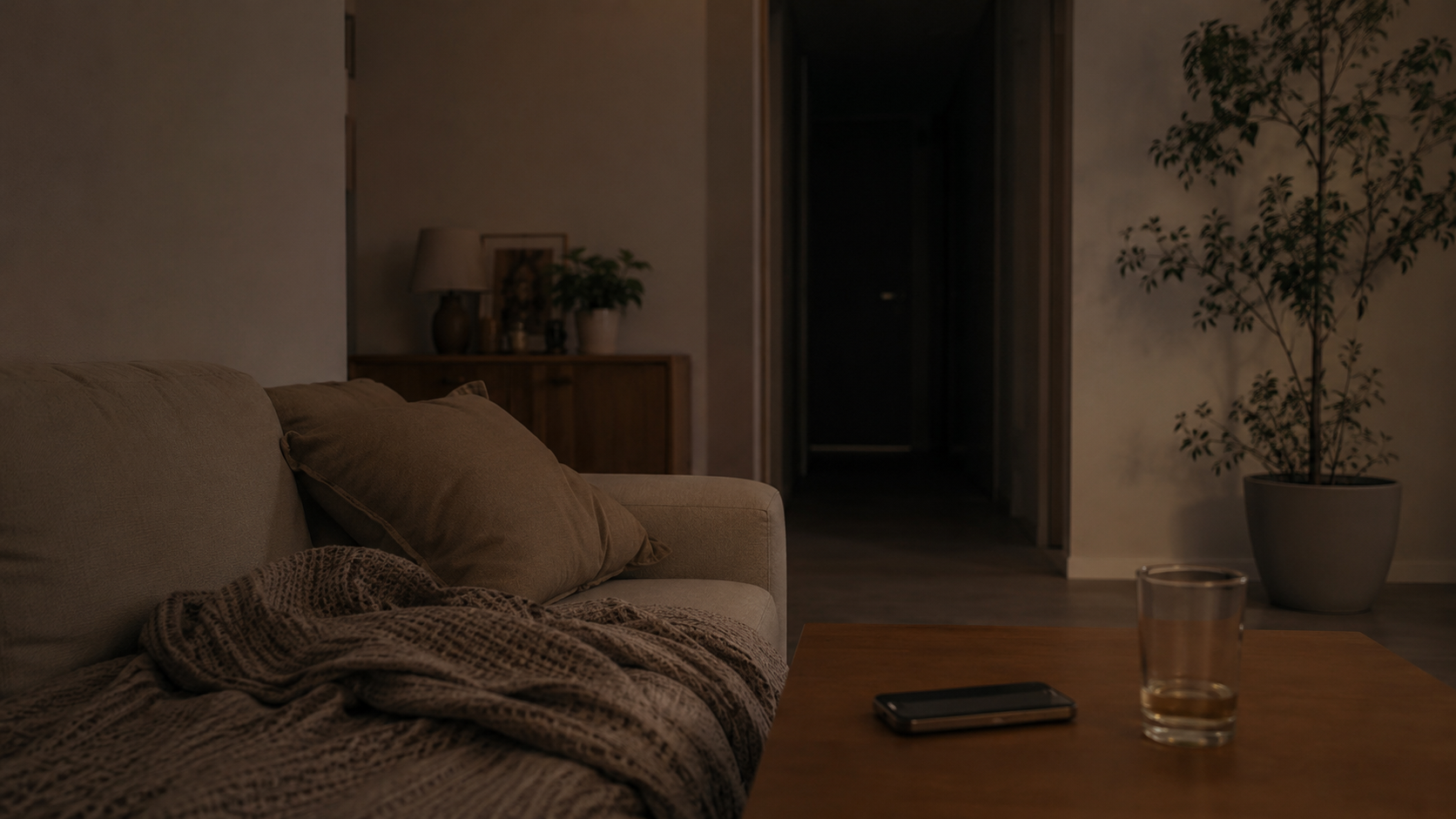 夜のリビング。ソファのブランケットとテーブルのスマホ。奥に洗面所の入口が暗いまま見えている
