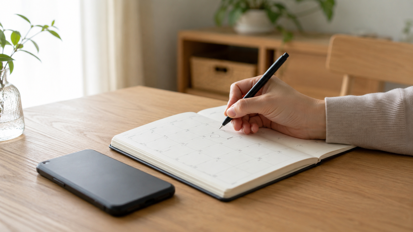 木のテーブルの上で、開いた手帳のカレンダーに予定を一つだけ書き込んでいる手元。ほかの日付は空いたまま。横にスマホが伏せて置かれている