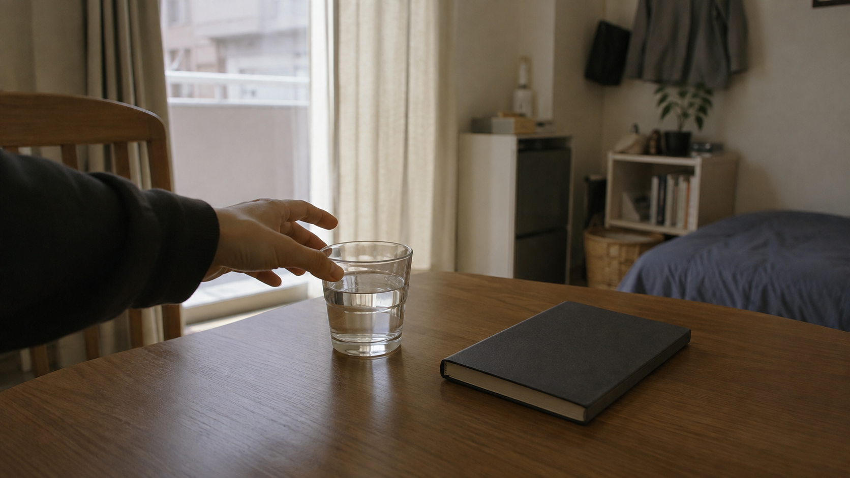 昼間の明るい部屋。木のテーブルに置いた水の入ったコップへ手を伸ばしているところ。横に閉じたままの手帳が一冊