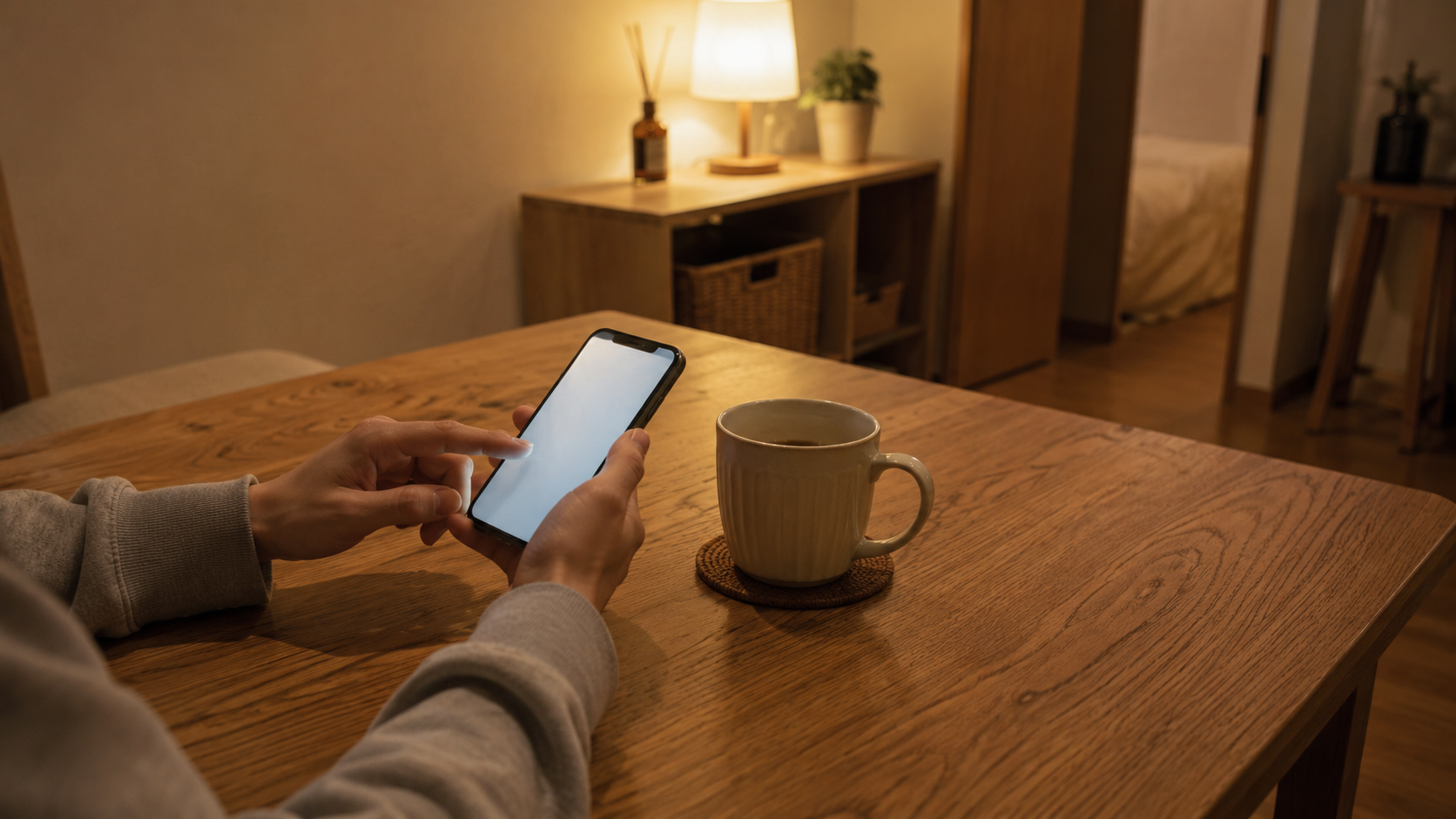 夜の部屋。木のテーブルでスマートフォンを操作している手元と、そばに置かれたマグカップ。画面の中身は読み取れない