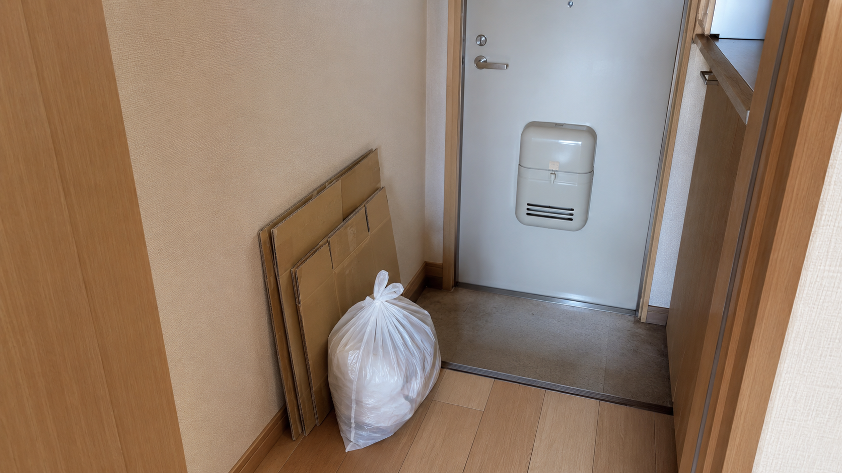 一人暮らしの玄関のそば。たたんだ段ボールが数枚壁に立てかけてあり、その横に口を結んだゴミ袋が一つ置かれている