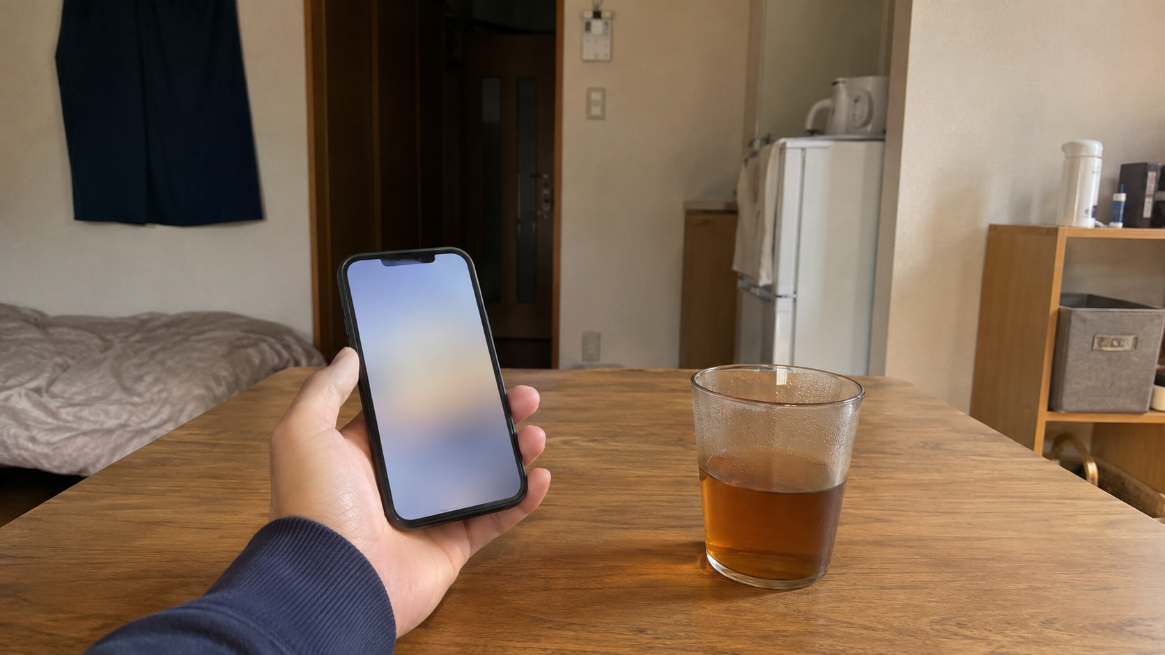 昼間の部屋。木のテーブルでスマートフォンを持っている手元と、横に置かれた飲み物の入ったコップ。画面の中身は読み取れない
