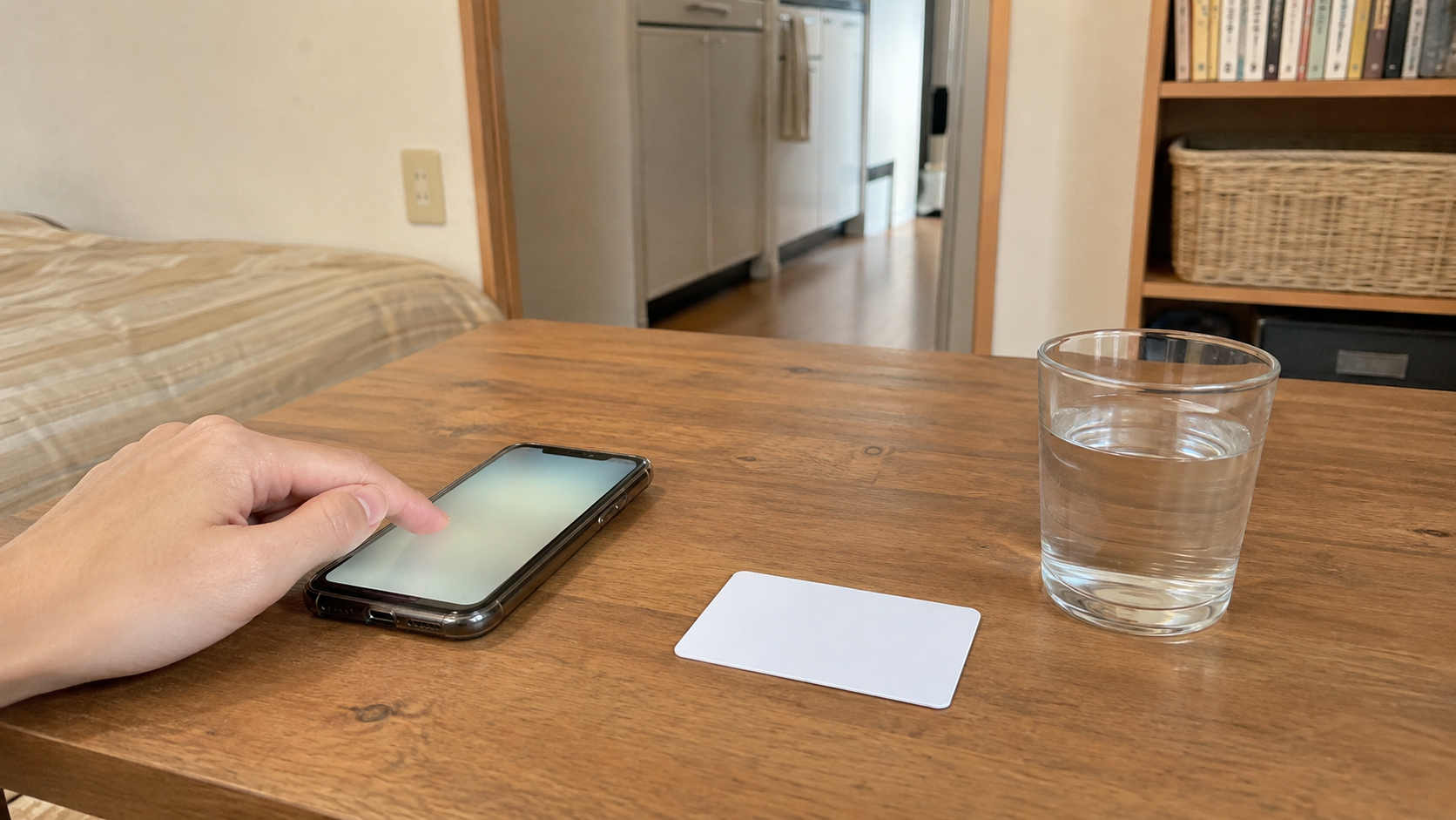 昼間の部屋。木のテーブルにスマートフォンと無地のカードが一枚、水の入ったコップが置かれ、スマートフォンに手を添えている